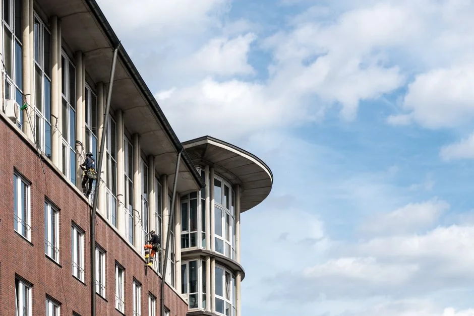 who-we-are Two workers clean windows on a modern glass building facade using a suspended platform.